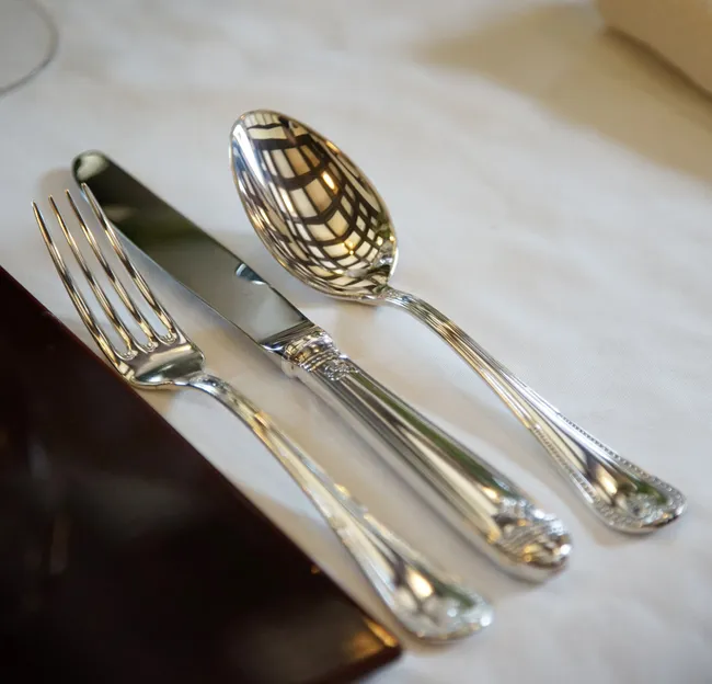 A fork, knife, and spoon with ornate handles neatly placed on a white tablecloth, with a napkin nearby and a ceiling reflection on the spoon.