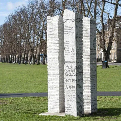 Tall stone sculpture engraved with text in multiple languages, standing on a grassy area outdoors.