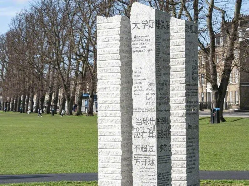 Tall stone sculpture engraved with text in multiple languages, standing on a grassy area outdoors.