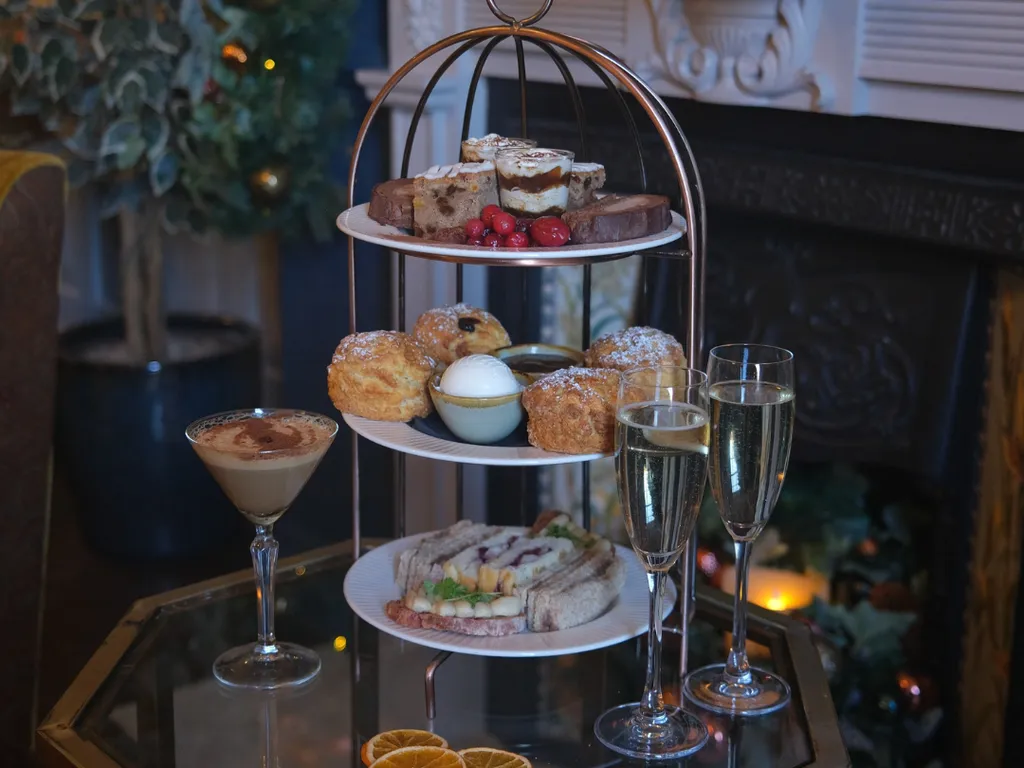 Three-tiered afternoon tea stand with assorted pastries, scones, and sandwiches, accompanied by two glasses of champagne and a cocktail on a glass table.