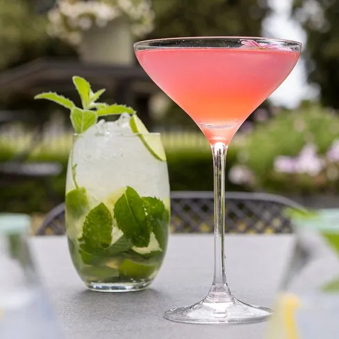 A pink cocktail in a martini glass next to a mojito garnished with mint leaves on an outdoor table.