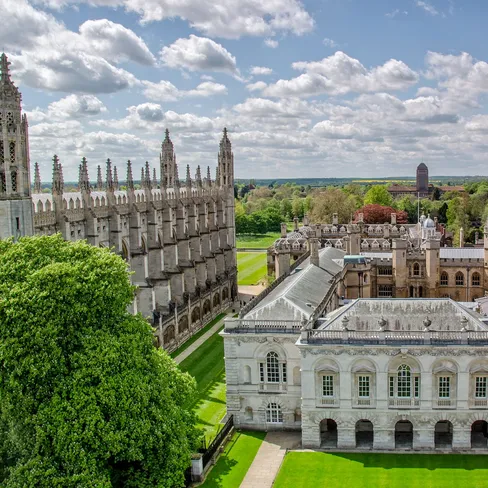 Aerial view of historic Cambridge University buildings with Gothic spires, lush green lawns, and trees under a partly cloudy sky.