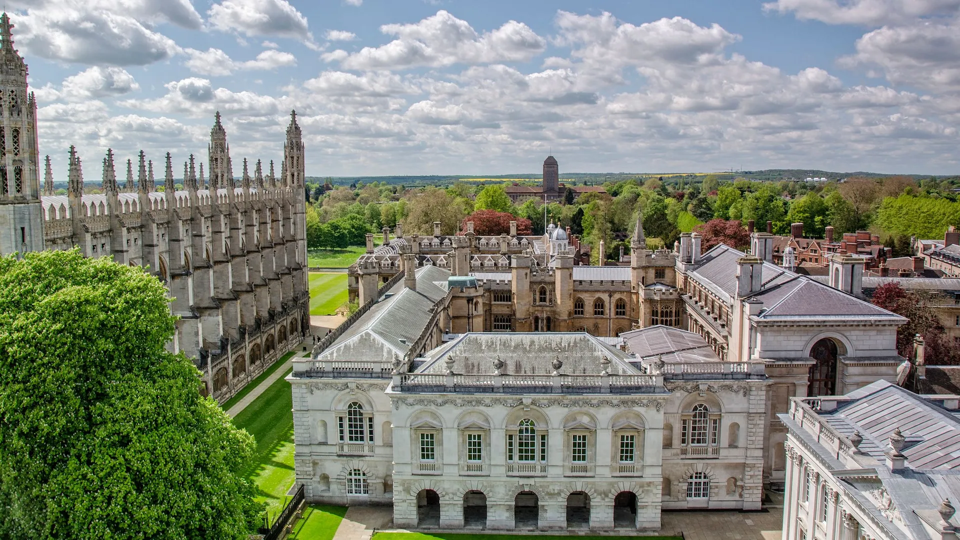 Aerial view of historic Cambridge University buildings with Gothic spires, lush green lawns, and trees under a partly cloudy sky.