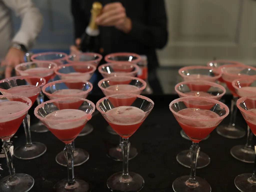 Several pink cocktails in sugar-rimmed martini glasses arranged on a tray, with people preparing drinks in the background.