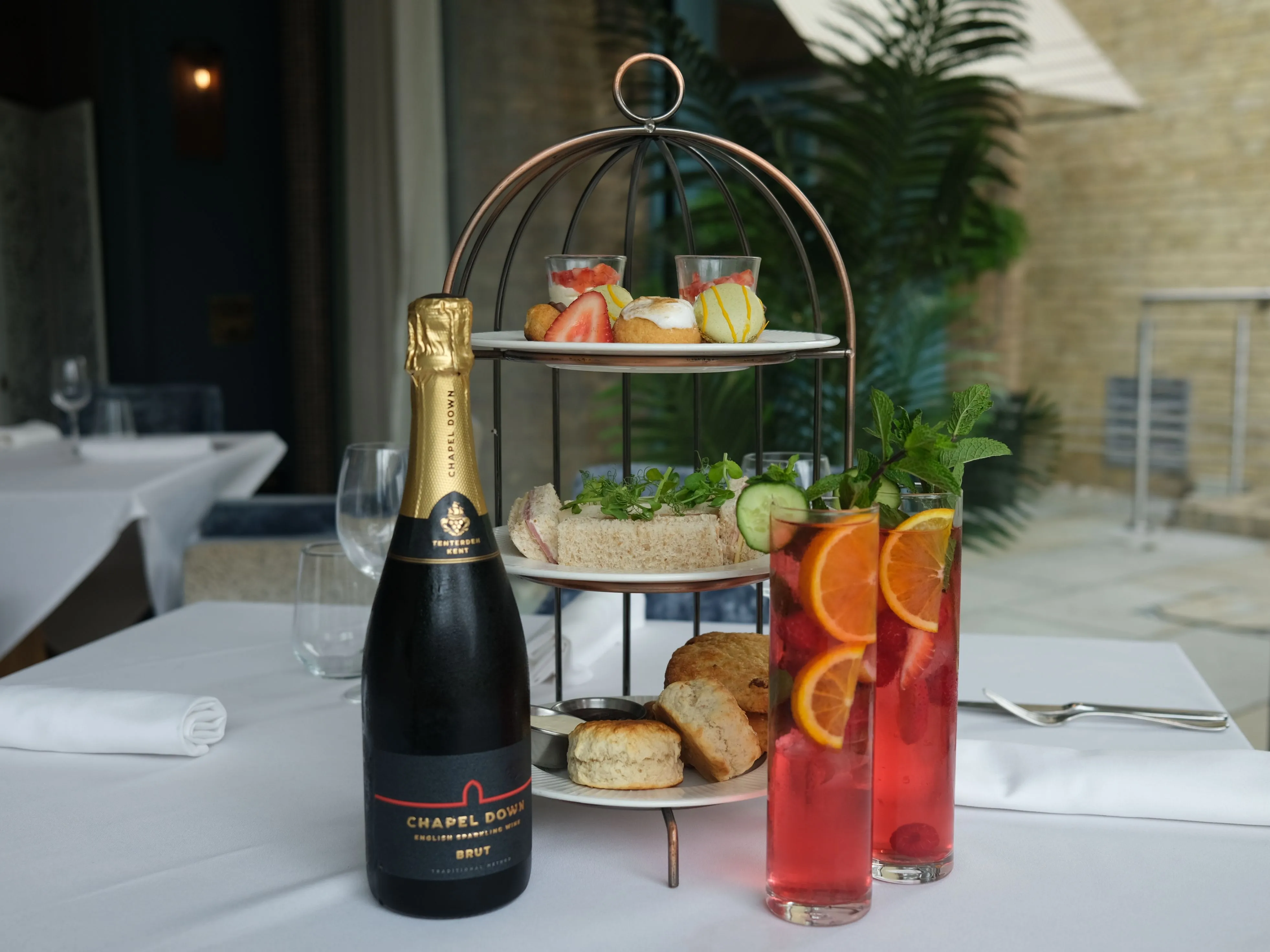 Afternoon tea set with sandwiches, scones, pastries, a bottle of sparkling wine, and two glasses of fruit cocktails on a restaurant table.