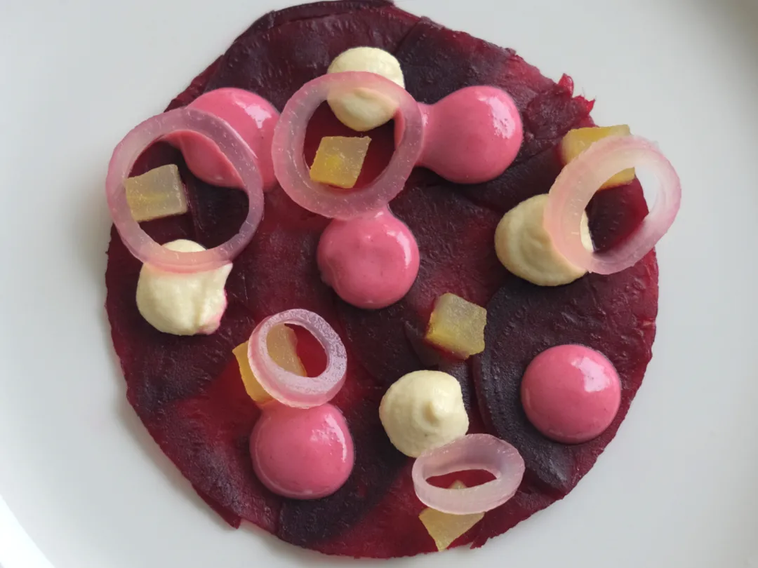 Artistic beetroot carpaccio garnished with dollops of mousse, pickled onion rings, and small cubes on a white plate