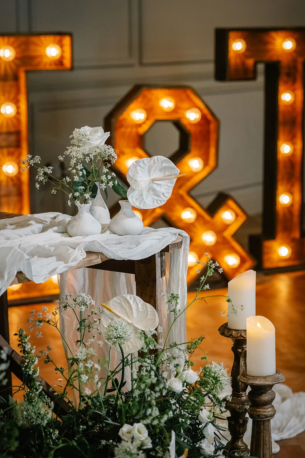 Elegant wedding-style floral display with white blooms, candles and illuminated ampersand and letter lights in the background
