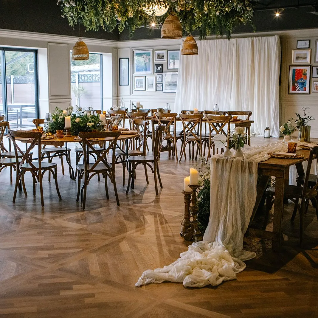 Stylish event space with wooden tables and chairs, draped fabric, candles and greenery hanging from the ceiling