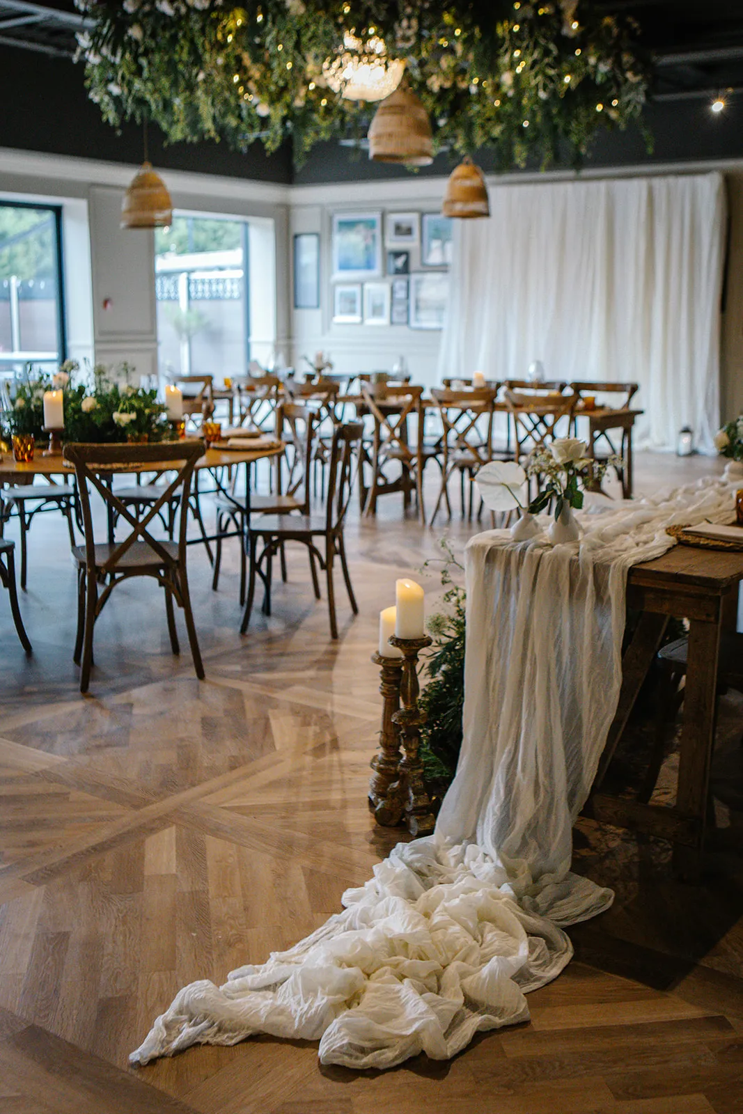 Rustic event space with wooden tables, cross-back chairs, candlelight and draped white fabric beneath hanging greenery