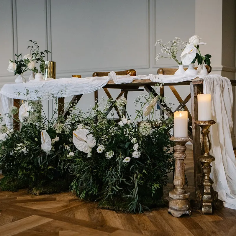 Elegant wedding sweetheart table with white draped linen, candles and lush white floral arrangements in a refined indoor venue