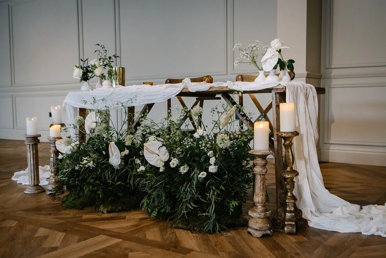 Elegant wedding sweetheart table with white draped linen, candles and lush white floral arrangements in a refined indoor venue