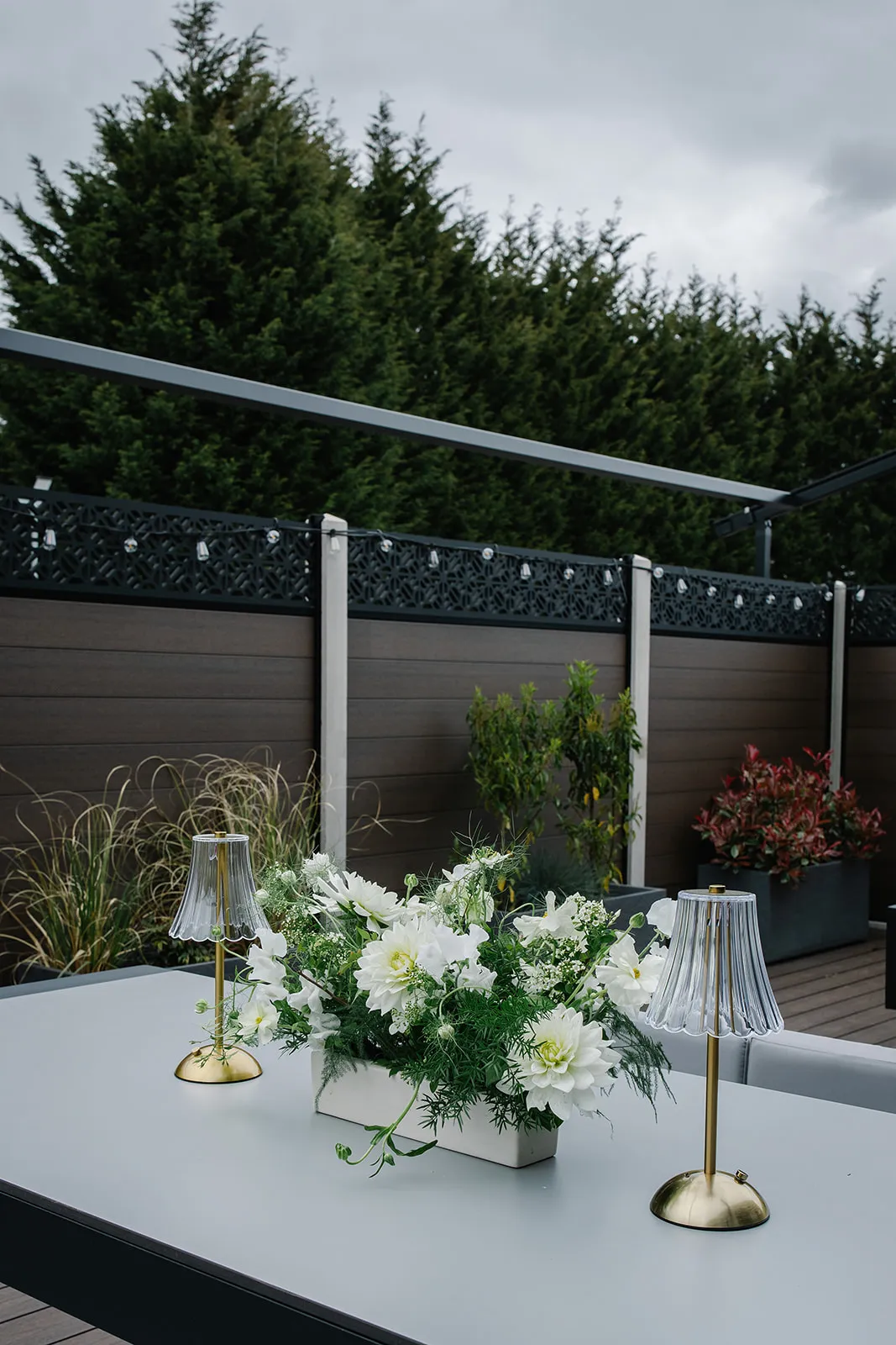 Outdoor hotel terrace table with white floral arrangement and two decorative lamps beside a fenced garden.