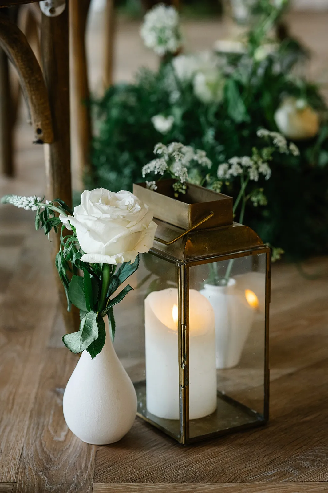 White rose in a small white vase beside a brass lantern with a lit candle on a wooden floor