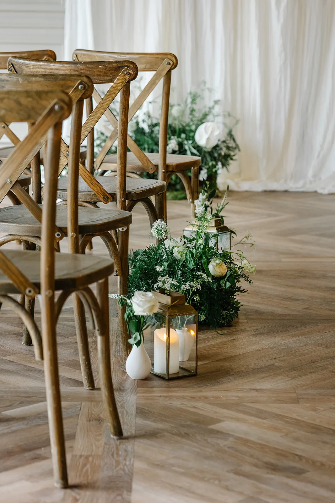 Rustic wooden chairs arranged for an indoor wedding ceremony with white flowers, greenery and lantern candles along the aisle.