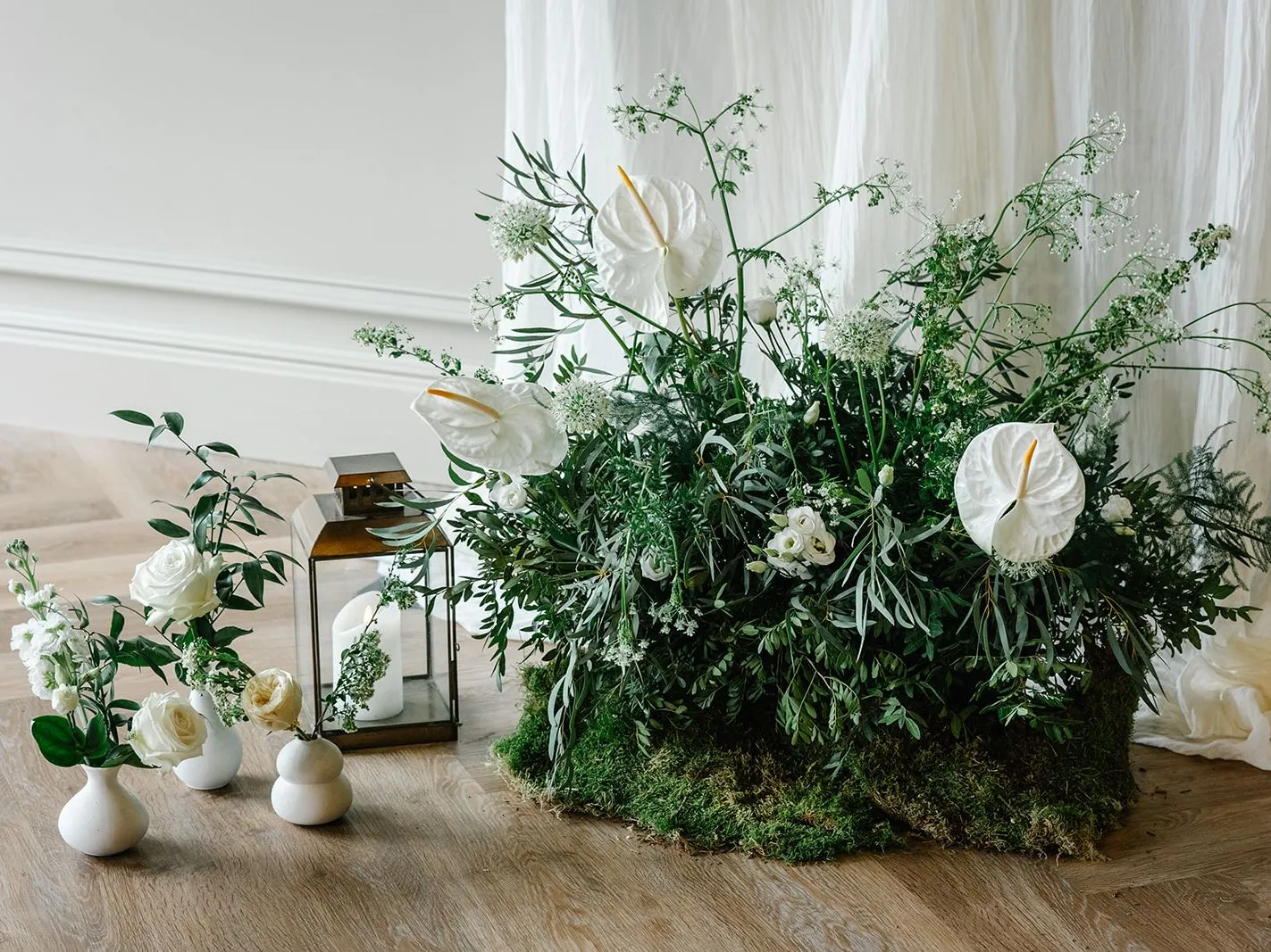 Elegant white floral arrangement with lush green foliage, small white vases and lanterns on a wooden floor against soft draped fabric.