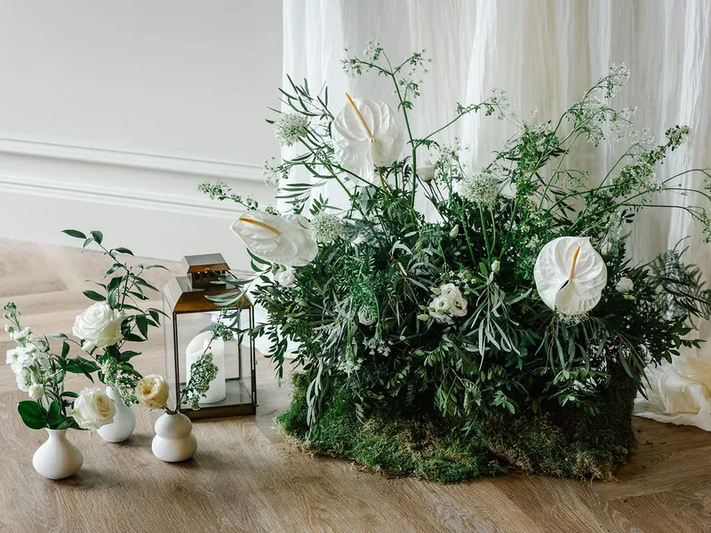 Elegant white floral arrangement with lush green foliage, small white vases and lanterns on a wooden floor against soft draped fabric.