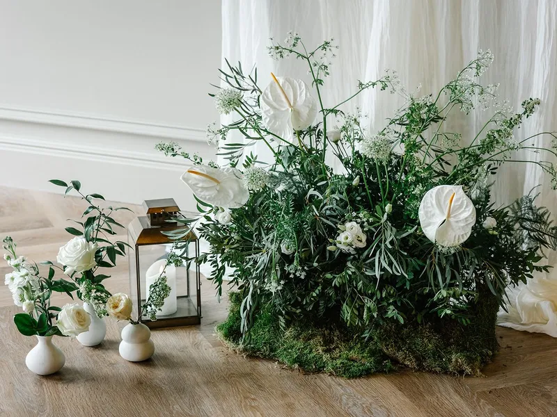 Elegant white floral arrangement with lush green foliage, small white vases and lanterns on a wooden floor against soft draped fabric.