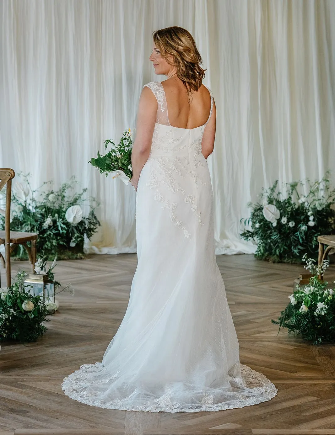 Bride in a fitted white wedding dress viewed from the back, holding a bouquet in an elegant indoor ceremony setting