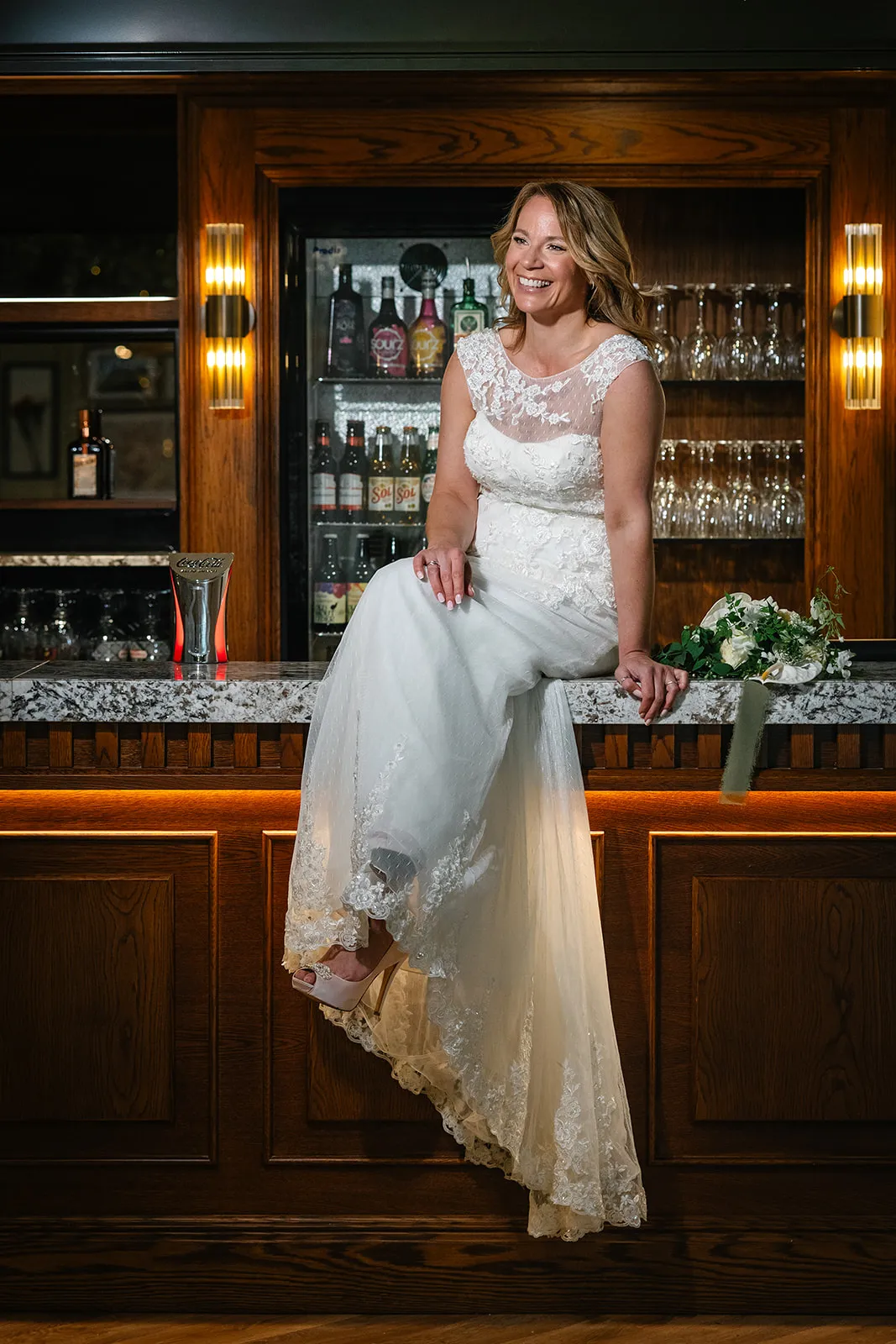 Bride in a lace wedding dress sitting on a wooden bar counter with her bouquet beside her, smiling in a warmly lit hotel bar.