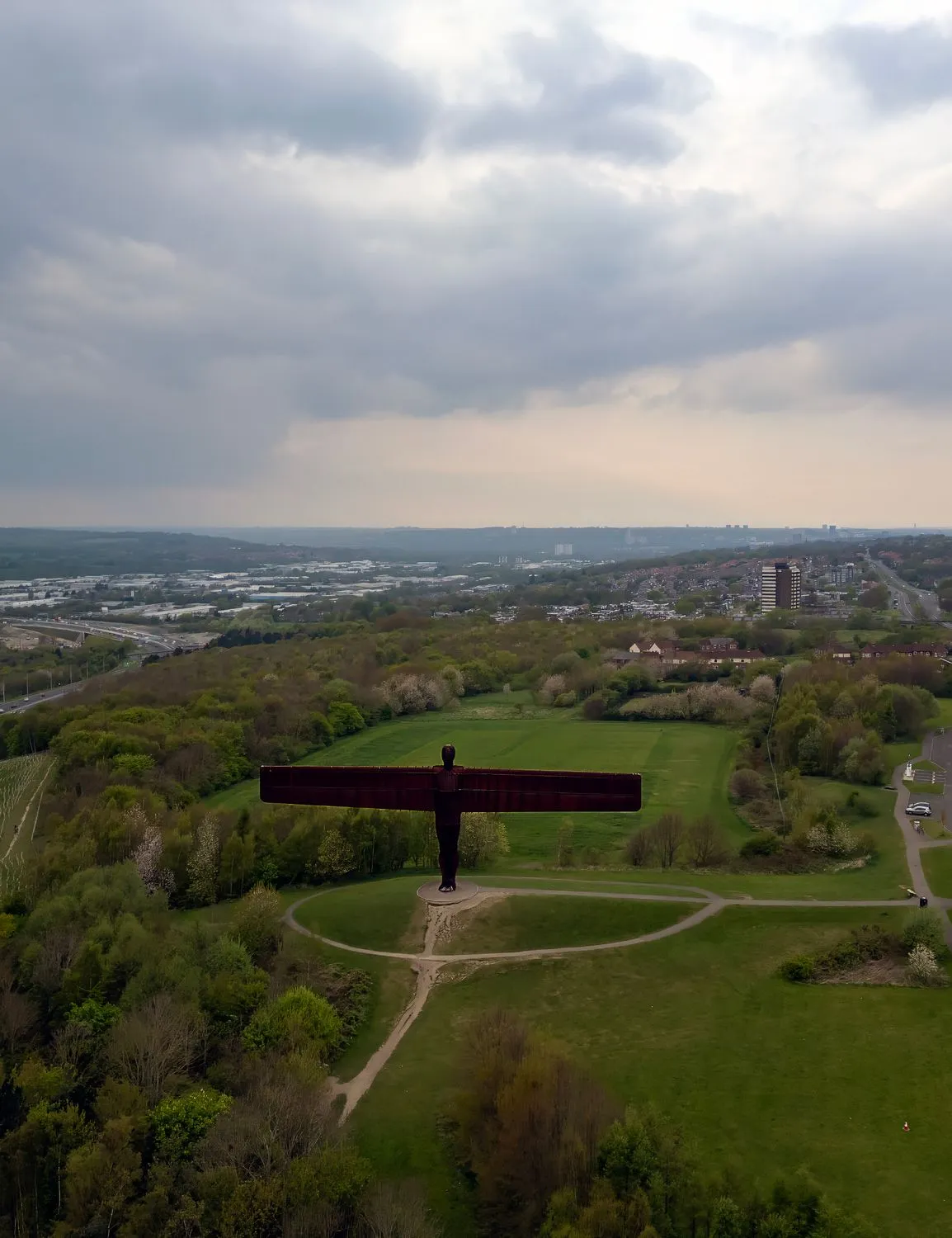 Aerial view of the Angel of the North standing in a grassy park beside major roads under dramatic cloudy skies.