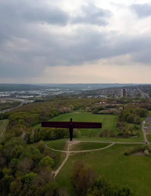 Aerial view of the Angel of the North standing in a grassy park beside major roads under dramatic cloudy skies.