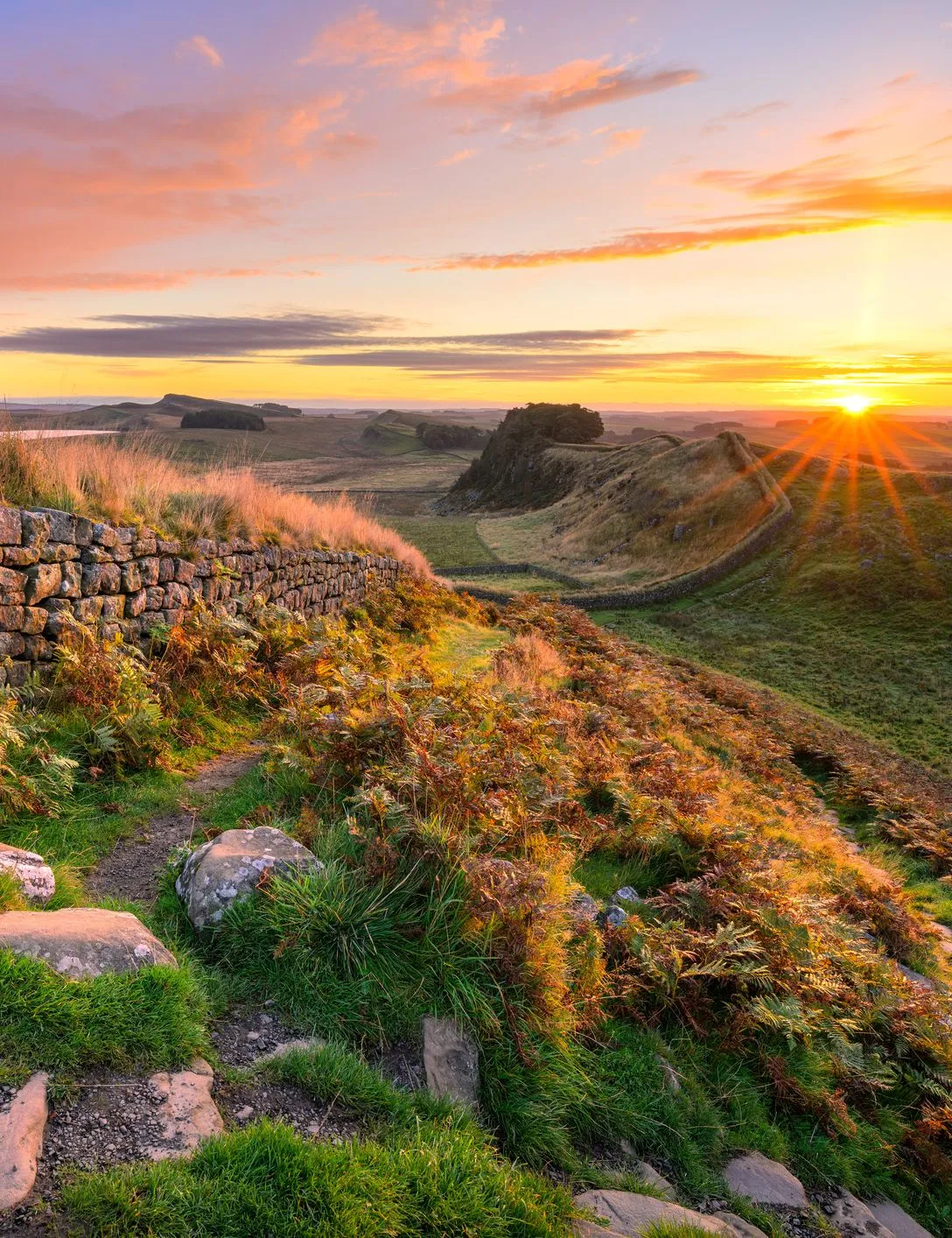 Sunrise over Hadrian's Wall with a stone path, grassy hillside and dramatic colourful sky.