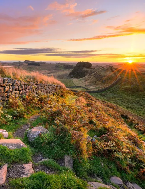 Sunrise over Hadrian's Wall with a stone path, grassy hillside and dramatic colourful sky.