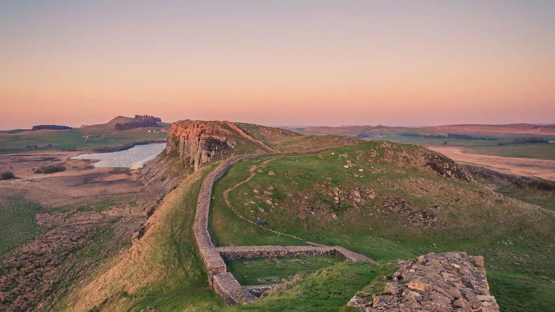 Hadrian's Wall winding over grassy hills at sunset under a soft pink and blue sky