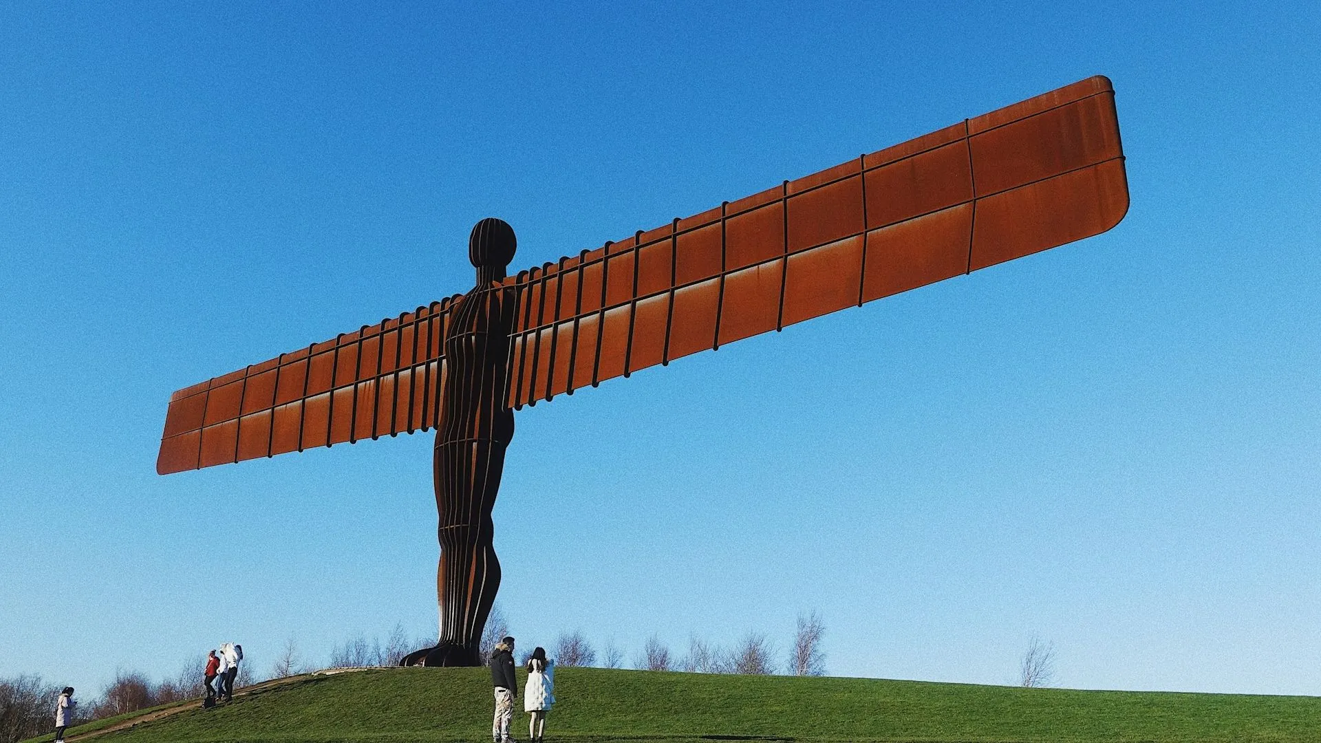 The Angel of the North sculpture standing on a grassy hill beneath a clear blue sky, with visitors nearby.