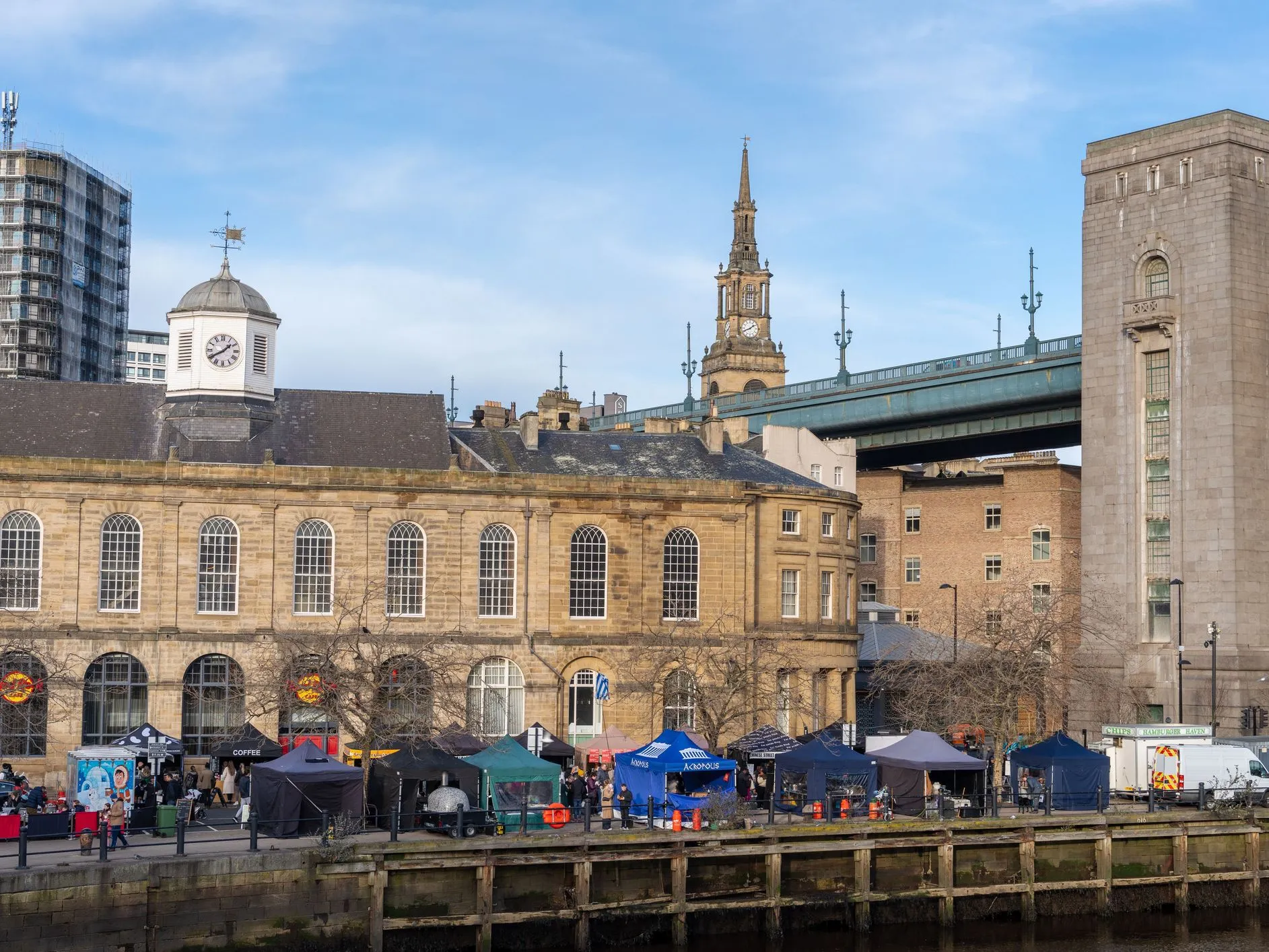 Quayside view of the Sandhill area in Newcastle upon Tyne, with historic stone buildings, market stalls and the Tyne Bridge above.