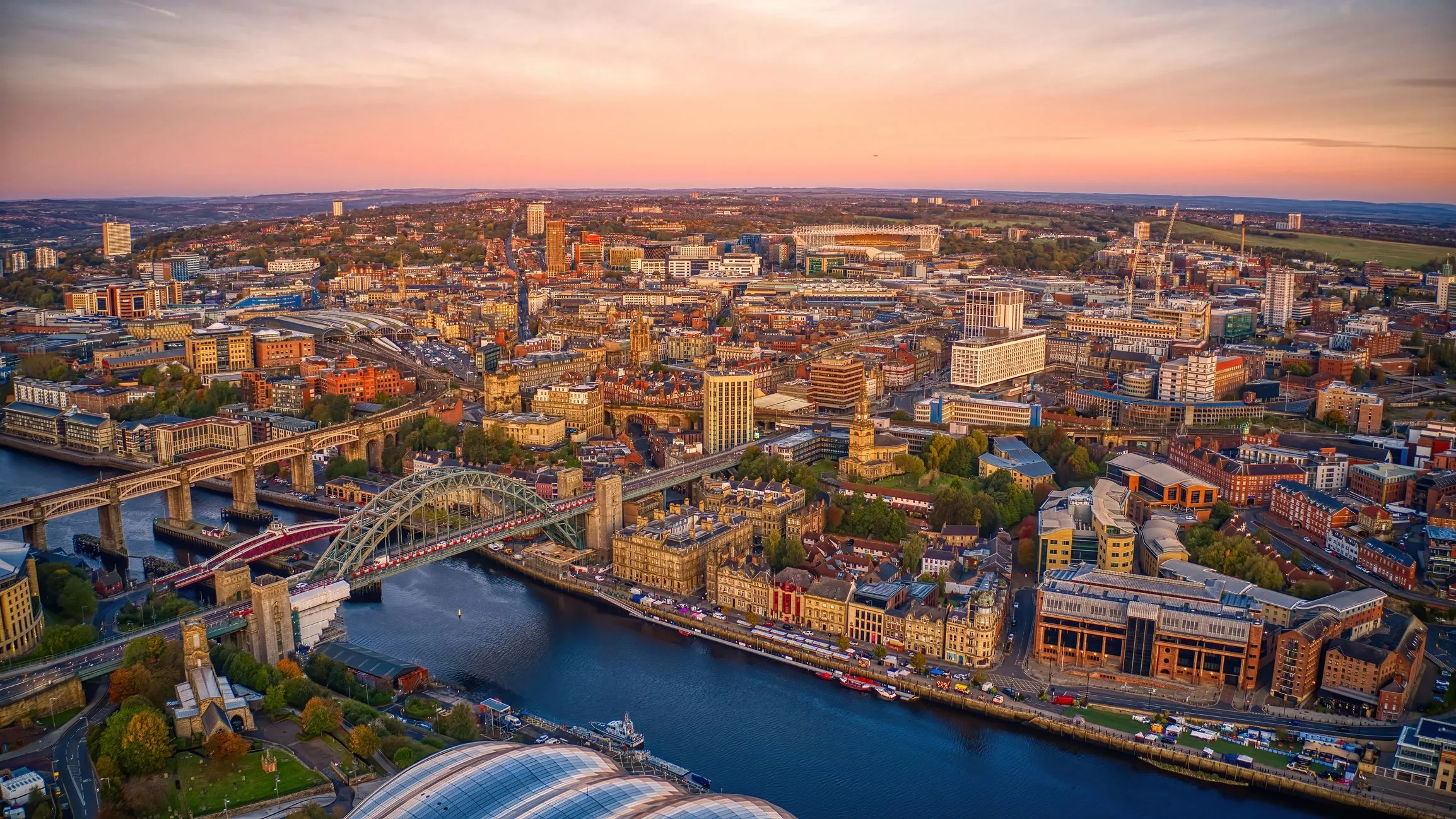 Aerial view of Newcastle upon Tyne at sunset, showing the River Tyne, the Tyne Bridge and the city skyline.