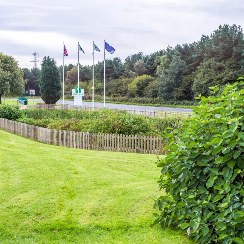 Exterior grounds at Holiday Inn Newcastle Gosforth Park with a neatly mown lawn, fenced garden and hotel sign by the roadside.