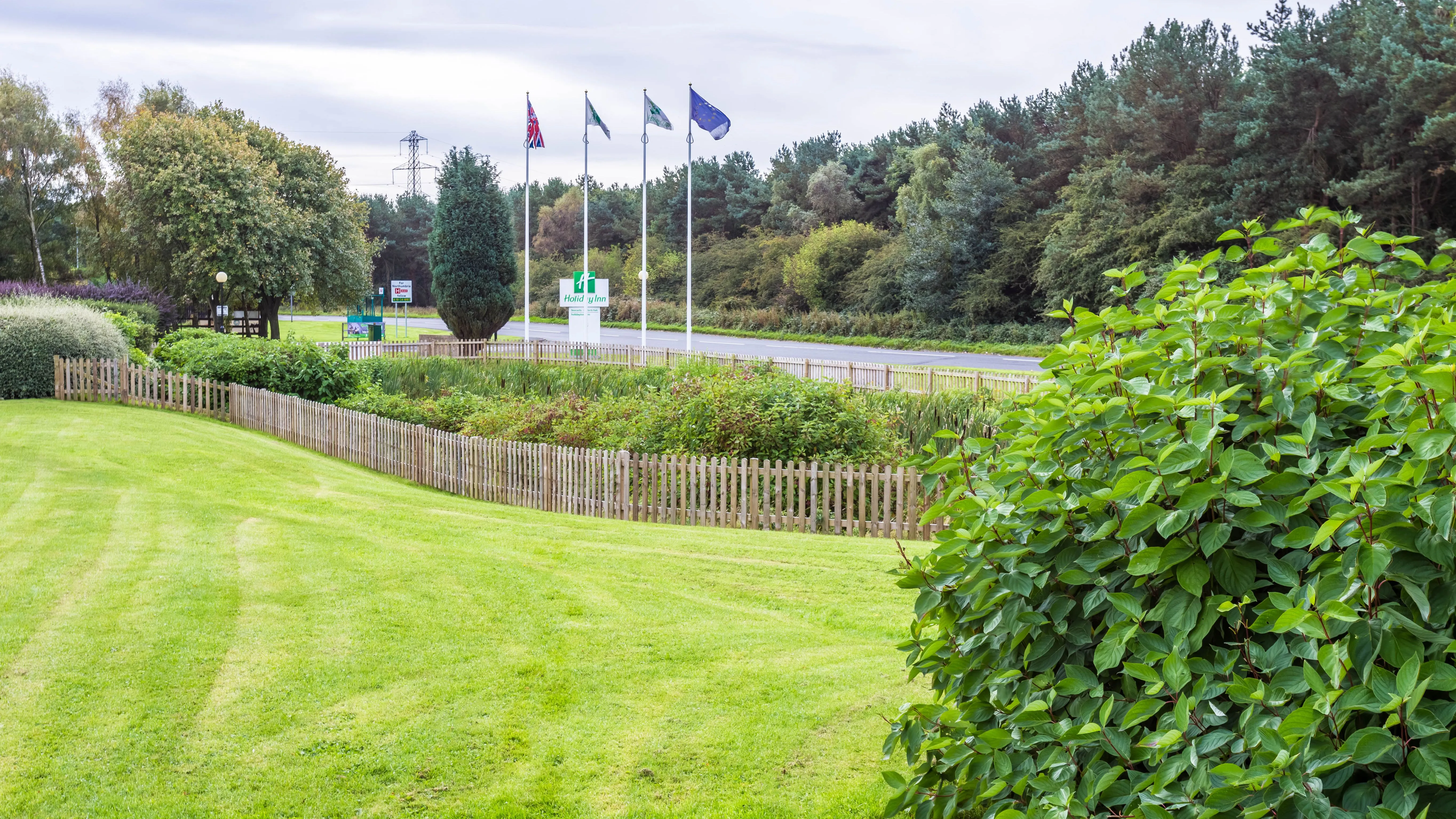 Exterior grounds at Holiday Inn Newcastle Gosforth Park with a neatly mown lawn, fenced garden and hotel sign by the roadside.
