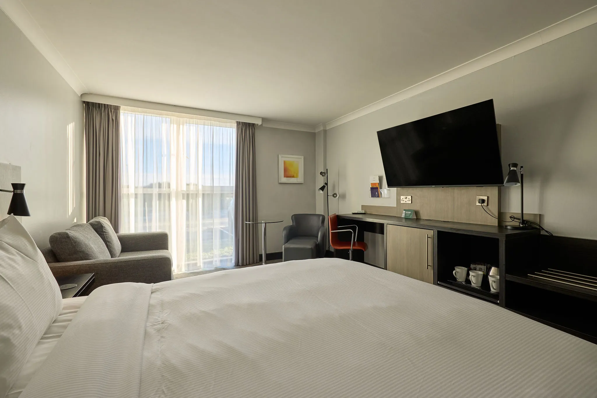 Modern hotel room with a large bed, wall-mounted television, desk, armchair and floor-to-ceiling window with sheer curtains.