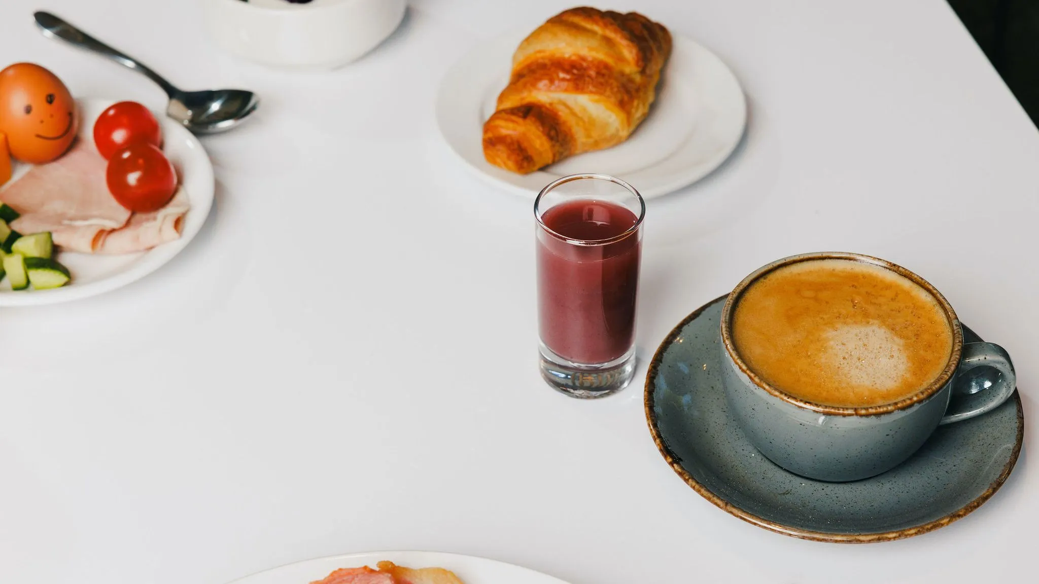 Breakfast spread with a cappuccino, croissant, yoghurt, juice, sliced ham, cucumber, tomatoes and eggs on a white table.