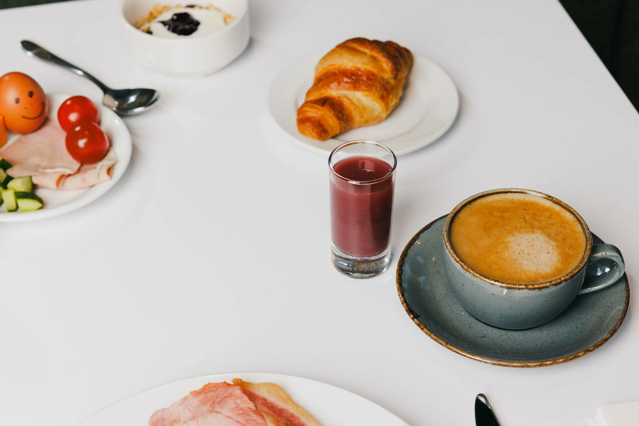 Breakfast spread with a cappuccino, croissant, yoghurt, juice, sliced ham, cucumber, tomatoes and eggs on a white table.