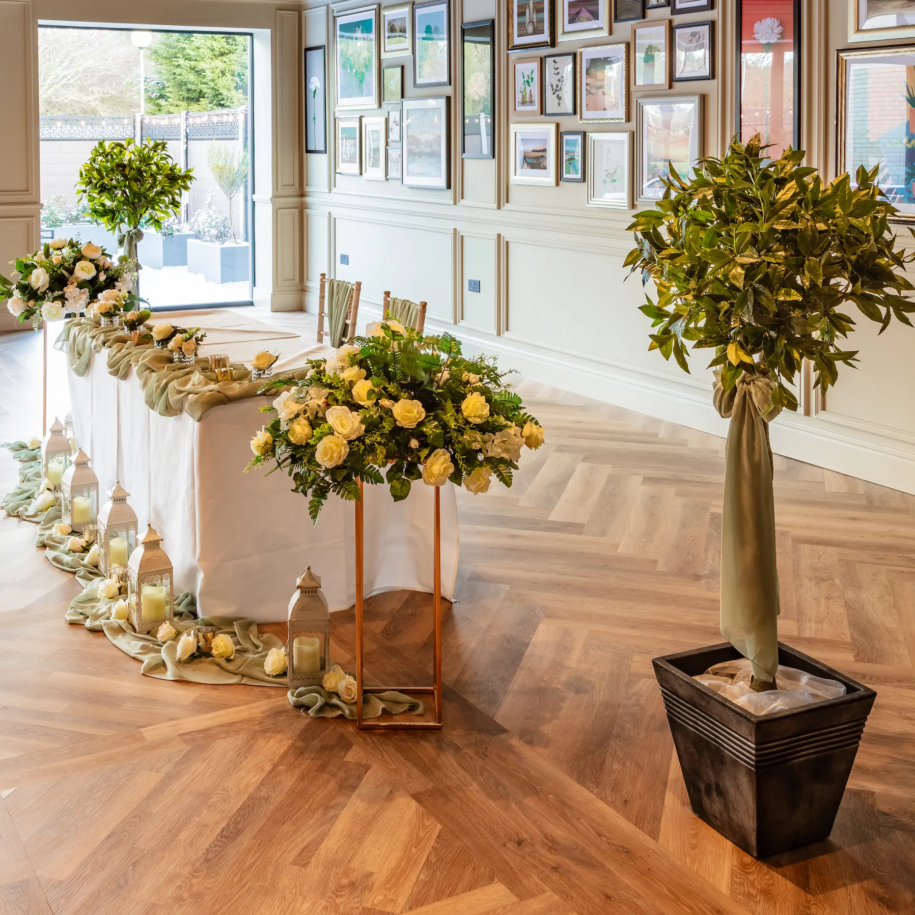 Elegantly styled wedding top table with white roses, greenery, lanterns and draped sage fabric in a bright hotel event room