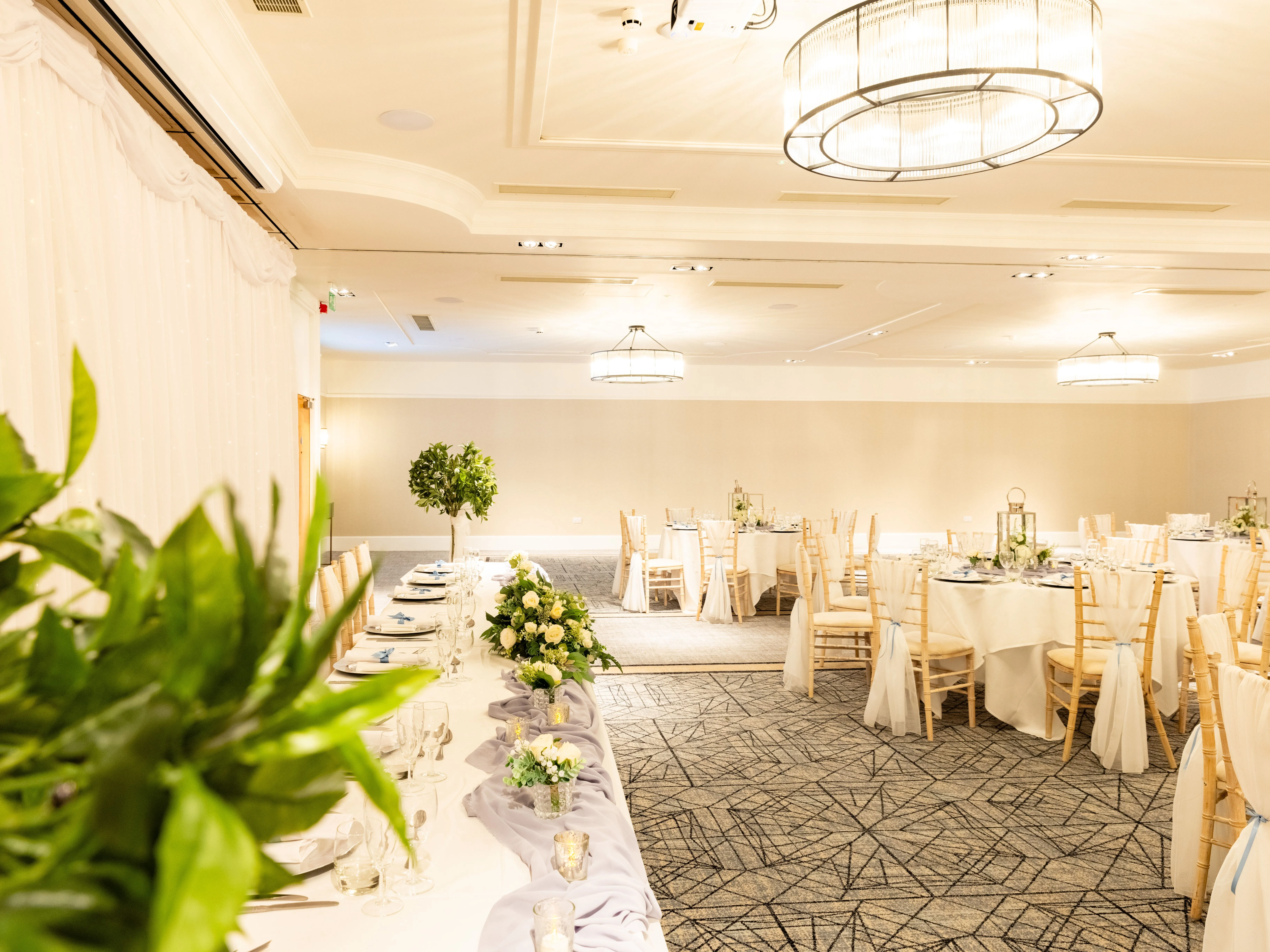 Elegantly decorated hotel function room set up for a wedding reception with round tables, floral centrepieces and draped backdrop