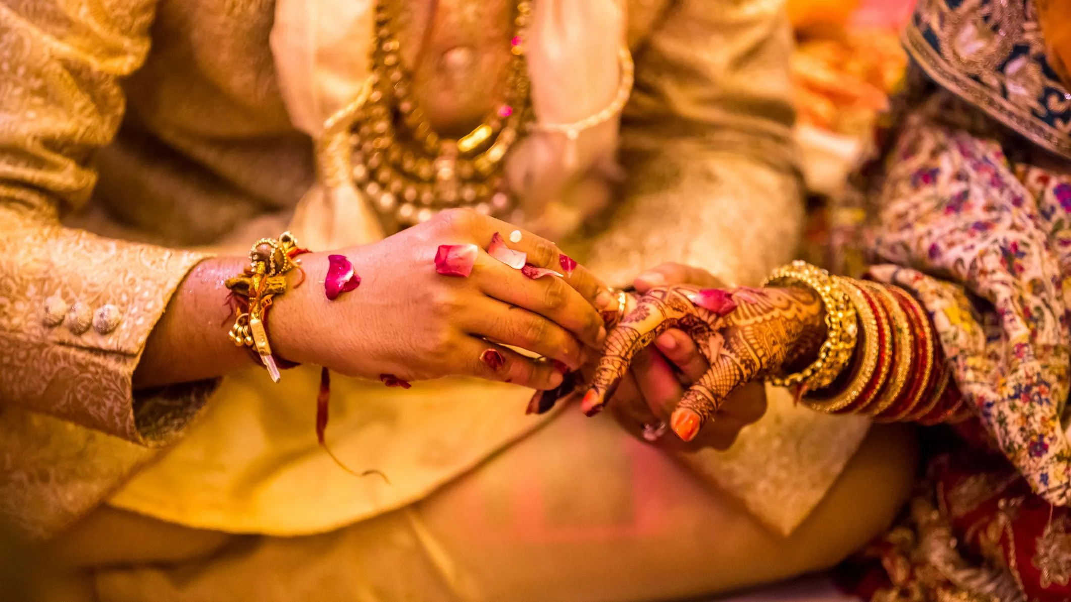 Close-up of a couple exchanging rings during a traditional South Asian wedding ceremony, with henna-decorated hands and rose petals.