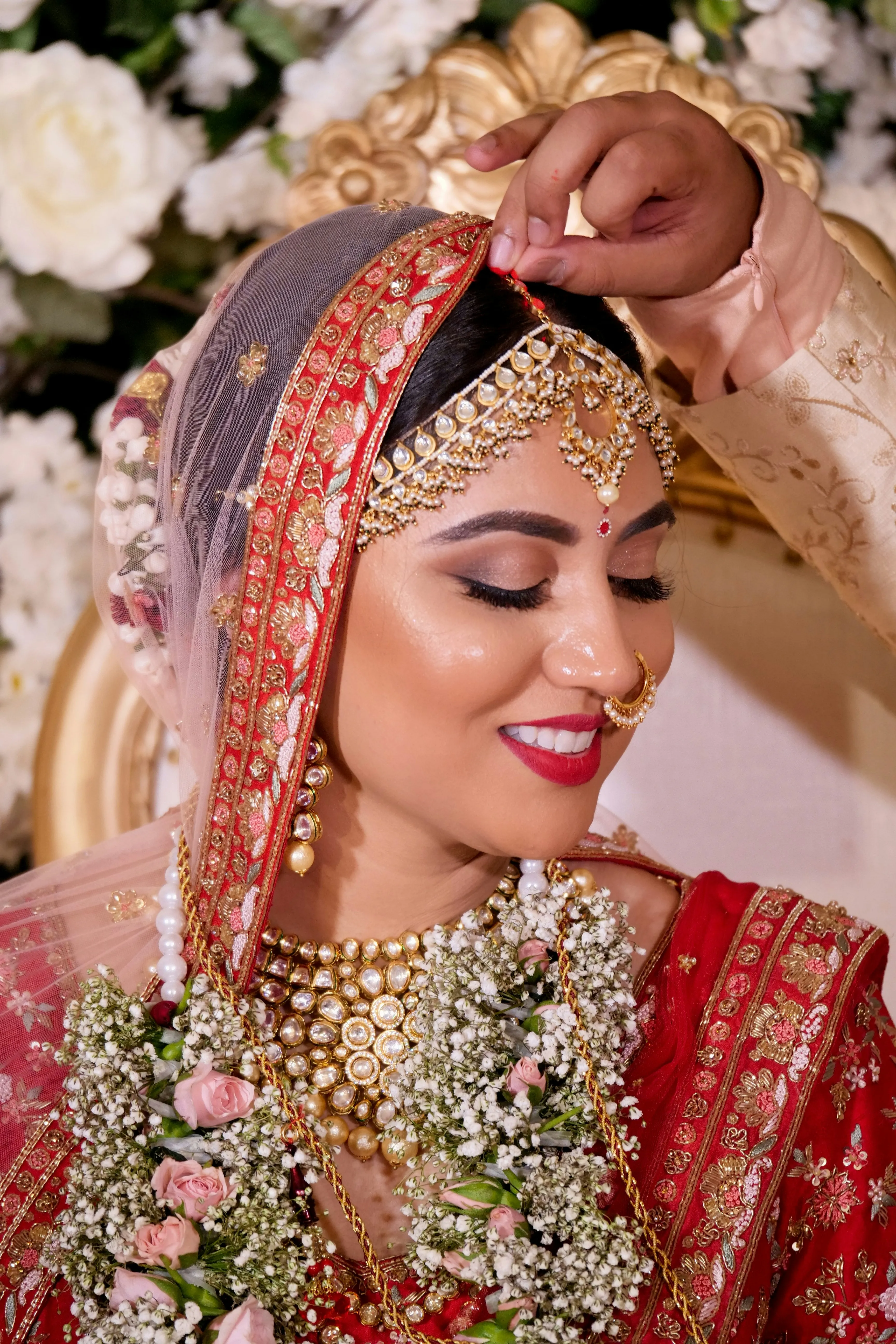 Close-up of a smiling bride in red and gold bridal attire as a hand adjusts her ornate head jewellery.