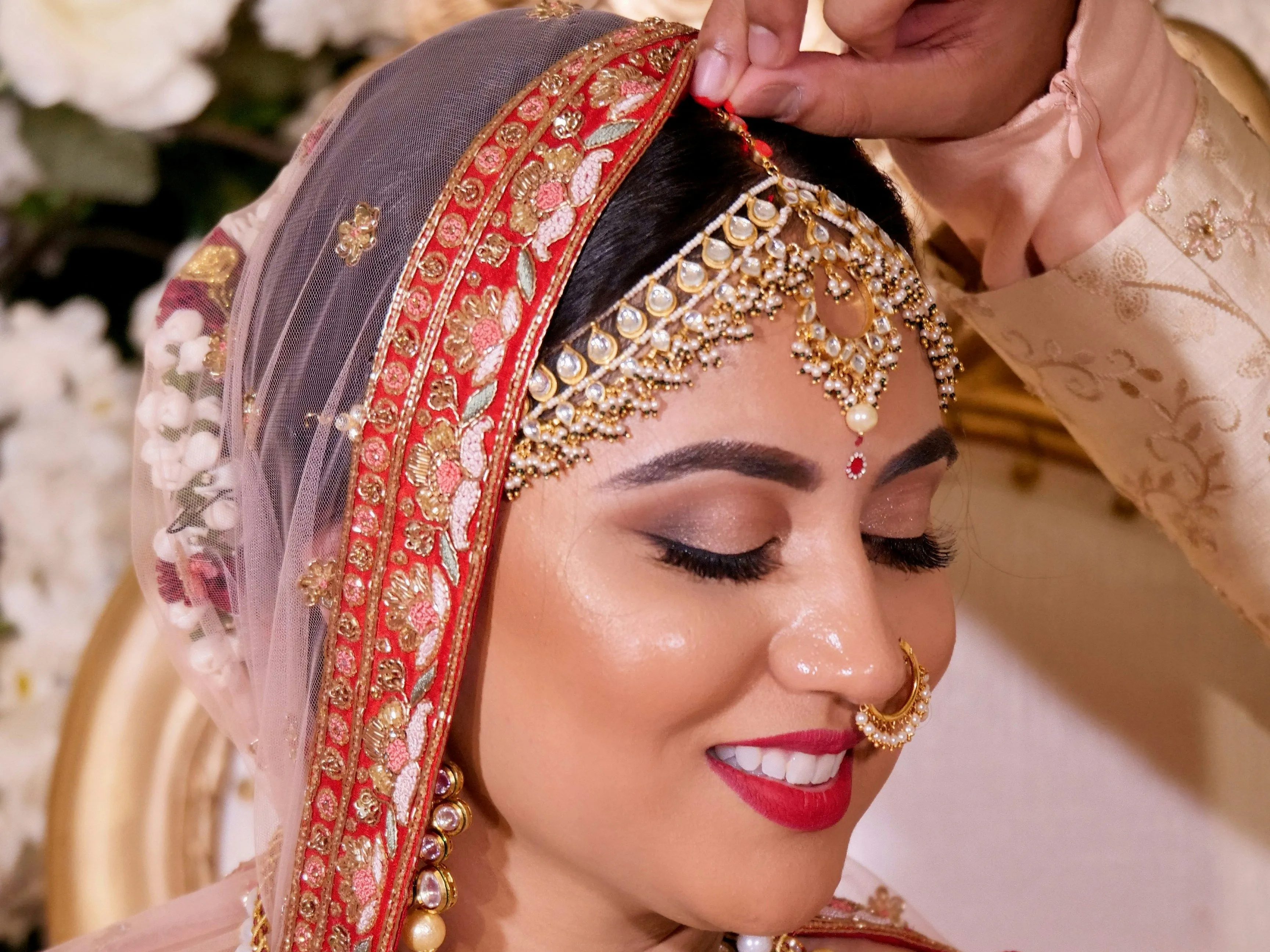 Close-up of a smiling bride in red and gold bridal attire as a hand adjusts her ornate head jewellery.