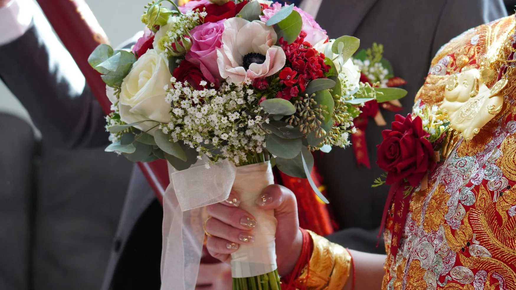 Close-up of a bride in ornate red and gold wedding attire holding a colourful bouquet of roses and mixed flowers.