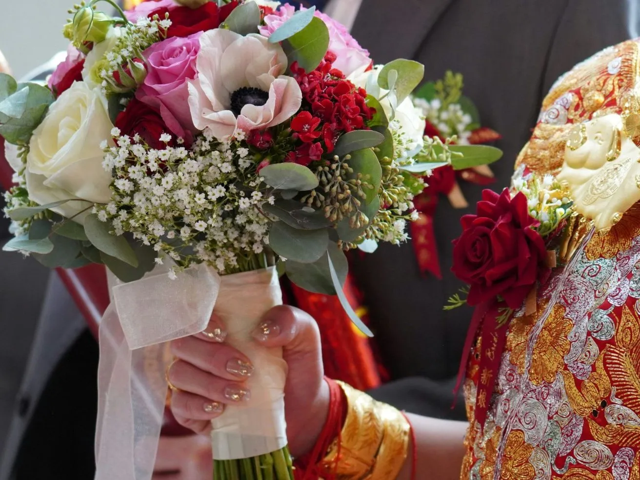 Close-up of a bride in ornate red and gold wedding attire holding a colourful bouquet of roses and mixed flowers.