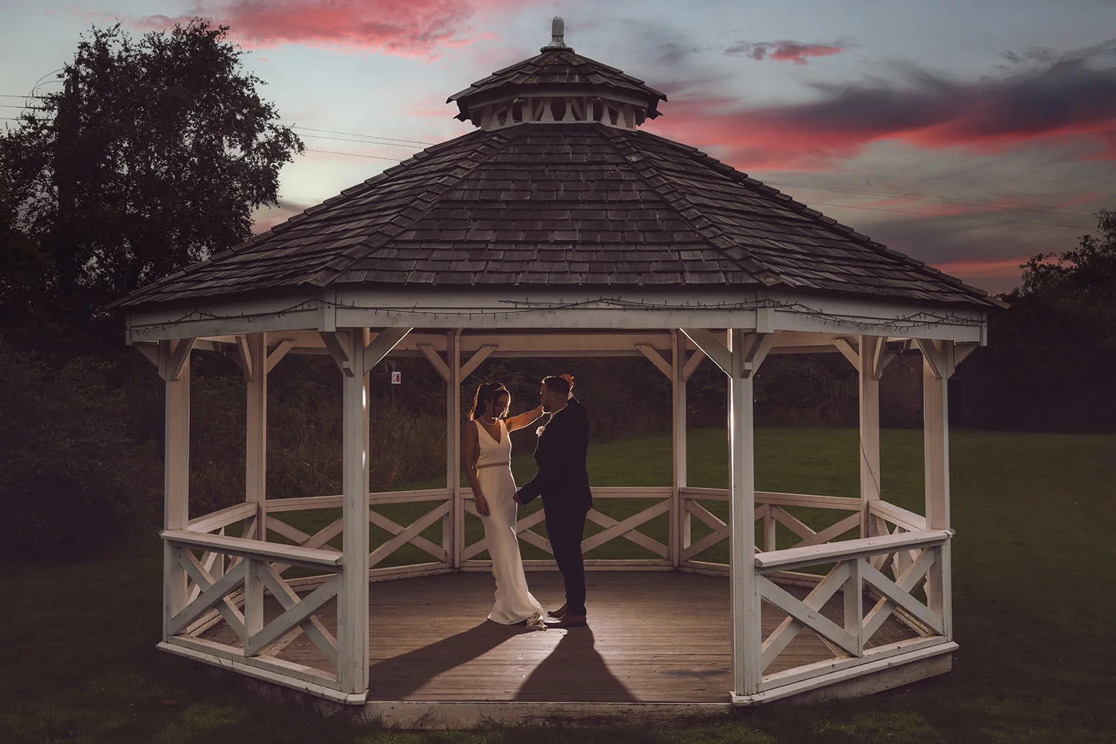 Bride and groom standing in a white wooden gazebo at sunset