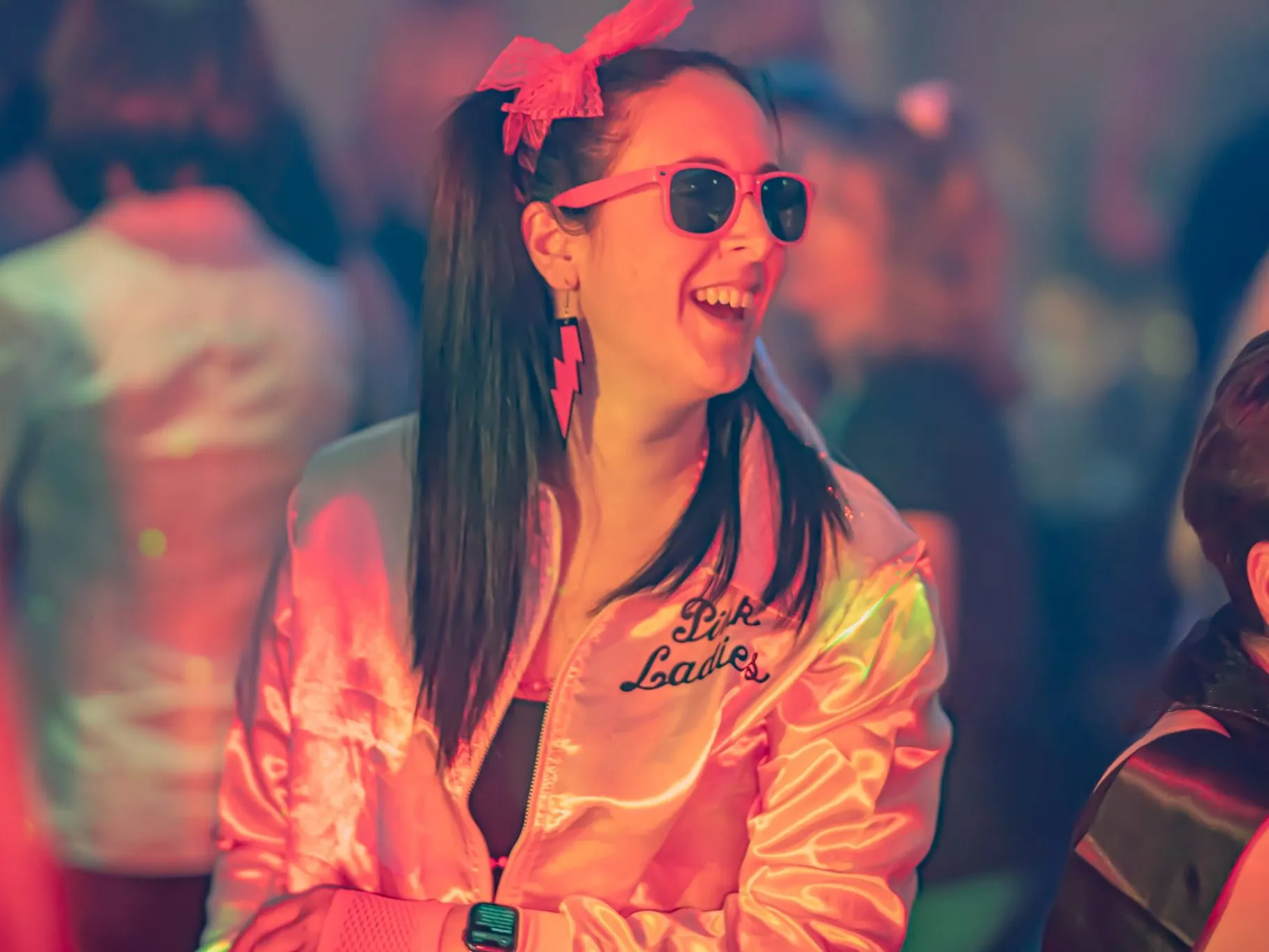 Woman in a pink satin jacket and pink sunglasses smiling at a lively party