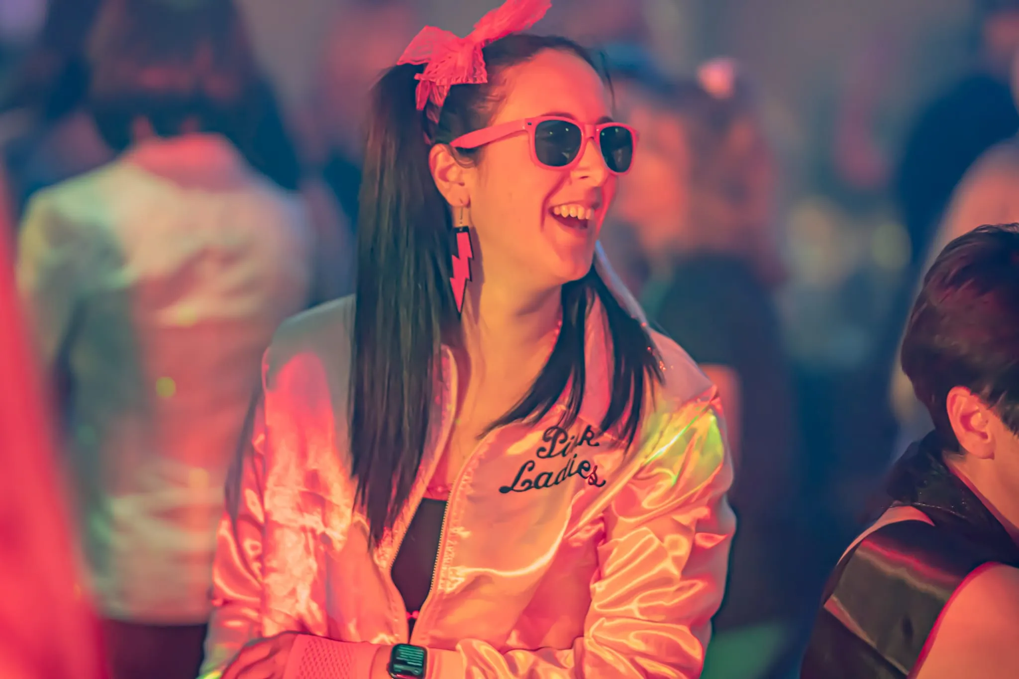 Woman in a pink satin jacket and pink sunglasses smiling at a lively party