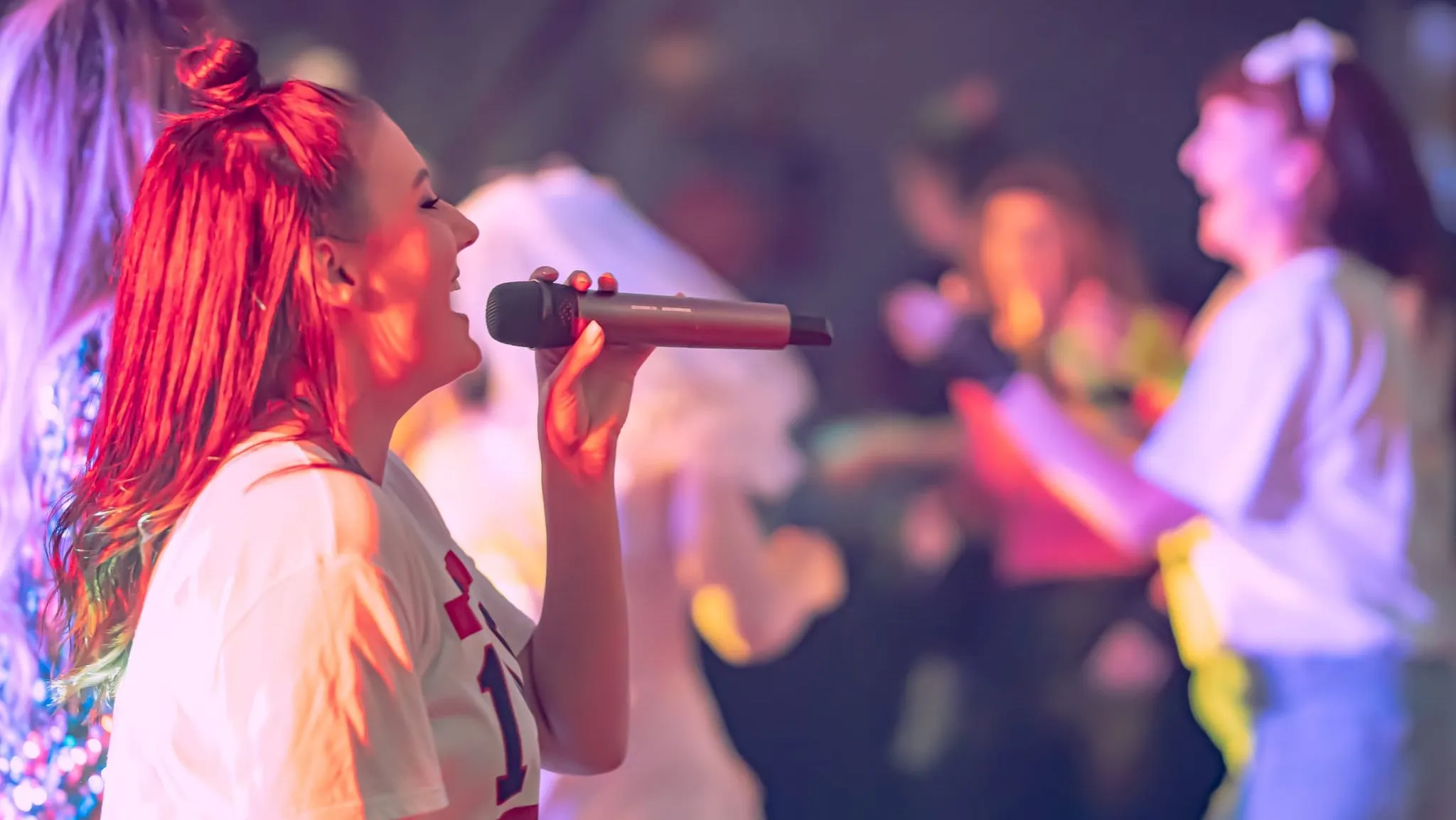 Woman singing into a microphone on stage under colourful lights, with blurred performers in the background.