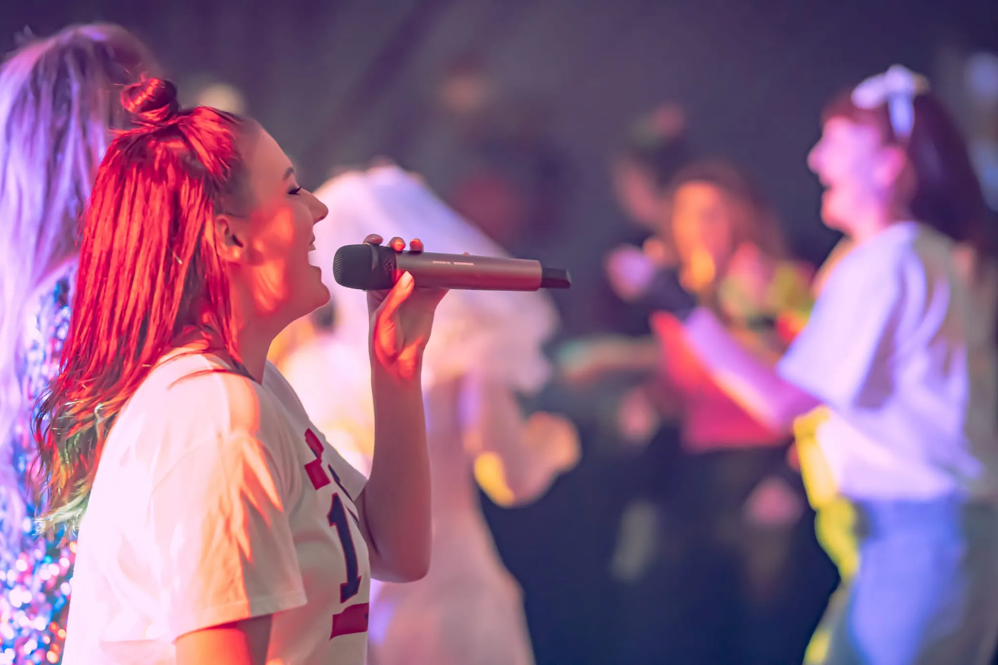 Woman singing into a microphone on stage under colourful lights, with blurred performers in the background.