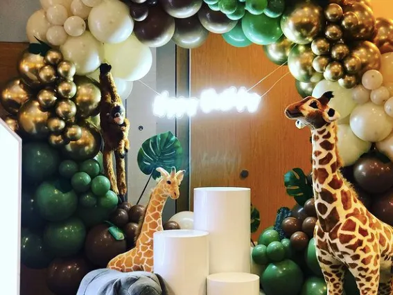Safari-themed balloon arch display with a giraffe, elephant and monkey around white plinths and tropical leaves.