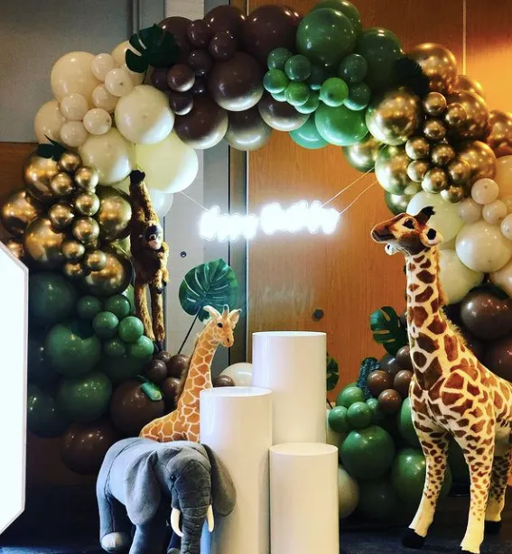 Safari-themed balloon arch display with a giraffe, elephant and monkey around white plinths and tropical leaves.